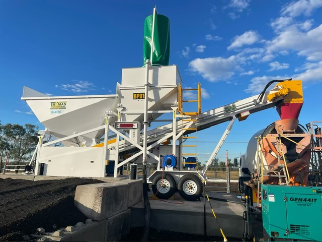A large mobile concrete batching plant with white and green components, a conveyor belt, and control equipment set up outdoors on a sunny day with a blue sky and scattered clouds.