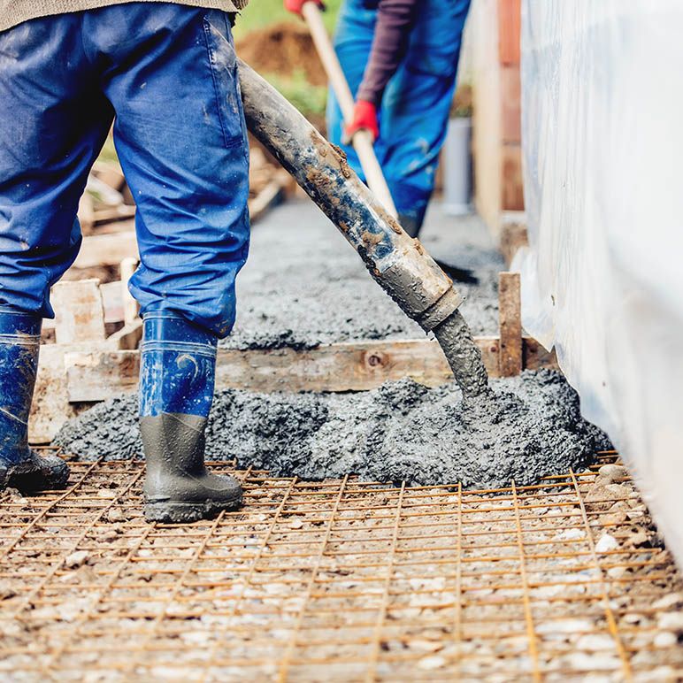 Construction workers pouring concrete onto a metal rebar grid, showcasing dynamic industry careers. One worker is holding a concrete hose while another operates in the background. Both are wearing blue pants and rubber boots, with their faces not visible in the image.