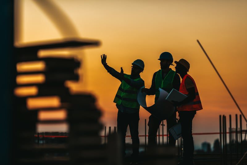 Three construction workers in high-visibility vests and hard hats stand on a building site at sunset. Two hold blueprints while the third gestures towards the structure, discussing the project. As they confer over their plans, the sunset provides a warm backdrop, silhouetting their figures. Contact Brawg for more details.