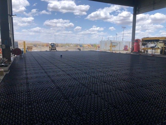 A large, industrial warehouse opens to a sunny outdoor area with construction equipment. The ground is covered with a grid-like, dark material. Solar panels and construction barriers are visible in the background under a partly cloudy sky.
