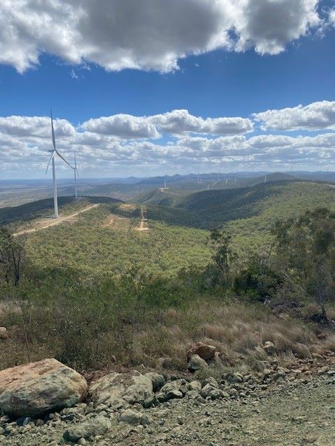 A scenic view of a hilly landscape with several wind turbines scattered across the area. The turbines stand tall above the green vegetation, under a partly cloudy sky. The foreground features rocky terrain, transitioning into lush greenery further away.