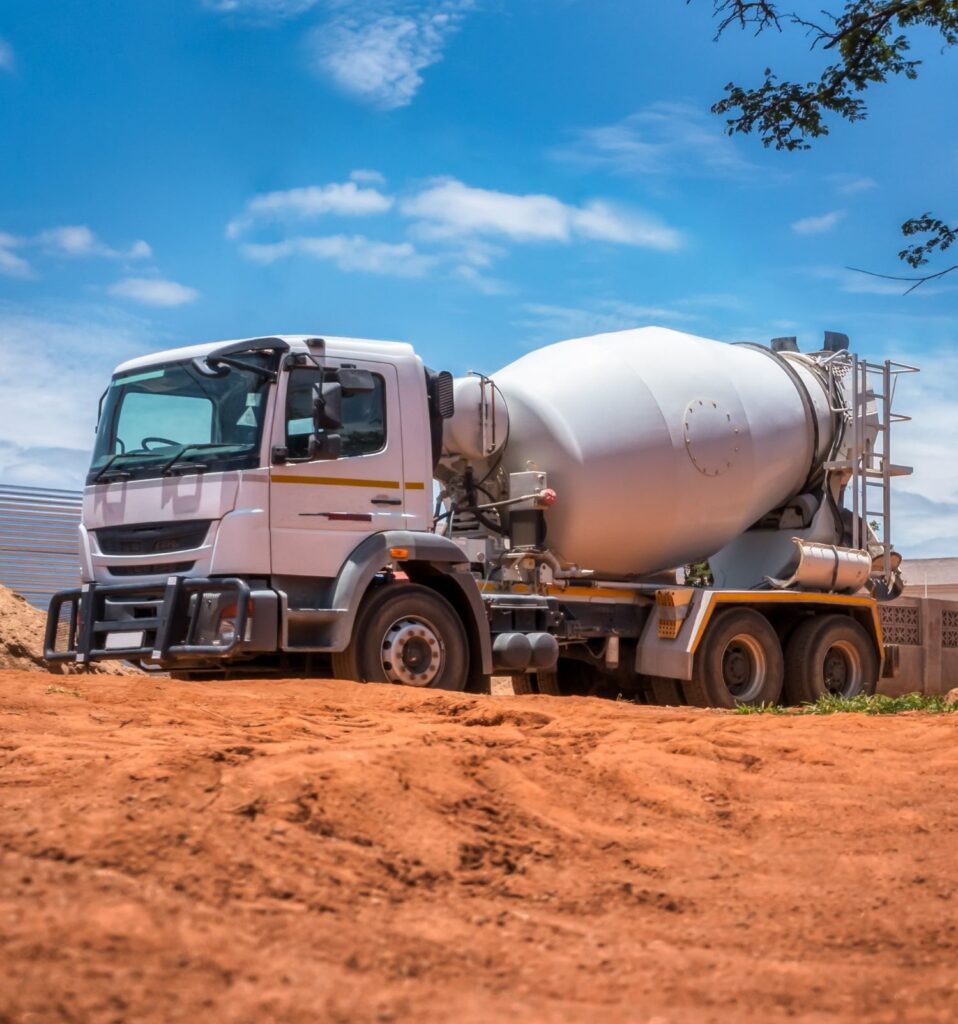 A white cement mixer truck with a large cylindrical drum is parked on a dirt construction site under a clear blue sky. The truck is facing left, and there are some construction materials and partial structures in the background.