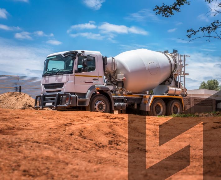 A white cement mixer truck is parked on a construction site with a dirt ground under a bright blue sky with scattered clouds. In the background, there are some piles of dirt, construction materials, and a partially visible building.