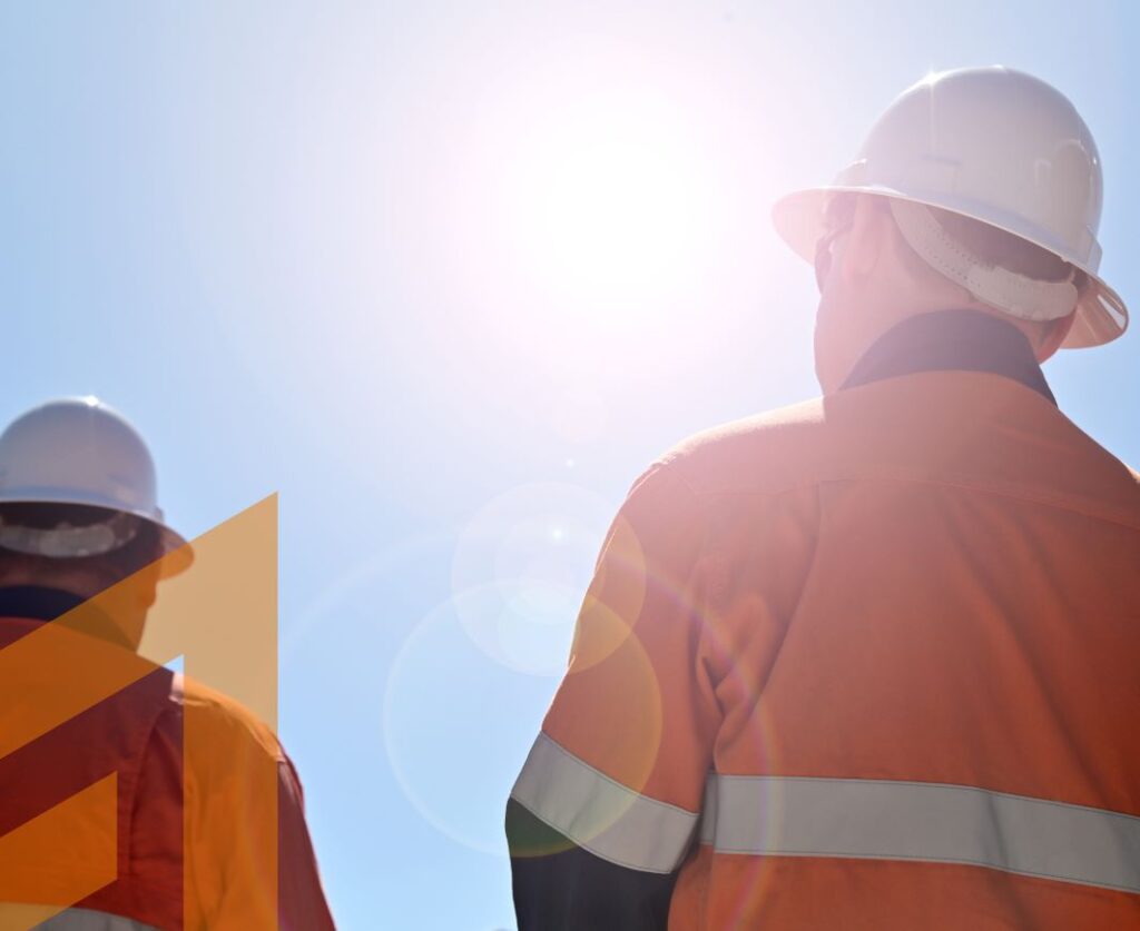 Two construction workers wearing white hard hats and orange safety vests with reflective strips stand outdoors under a bright sunlit sky. One worker faces away from the camera, while the other is partially visible. The sunlight creates a lens flare effect.