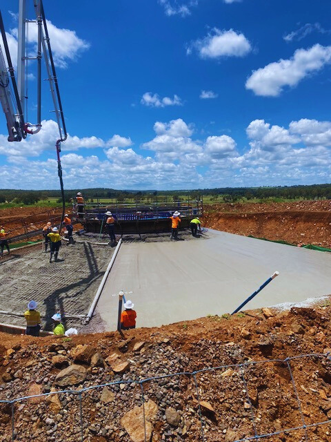 Construction workers in safety gear pour and smooth concrete on a large foundation slab at a construction site under a bright blue sky with scattered clouds and surrounding green landscape.