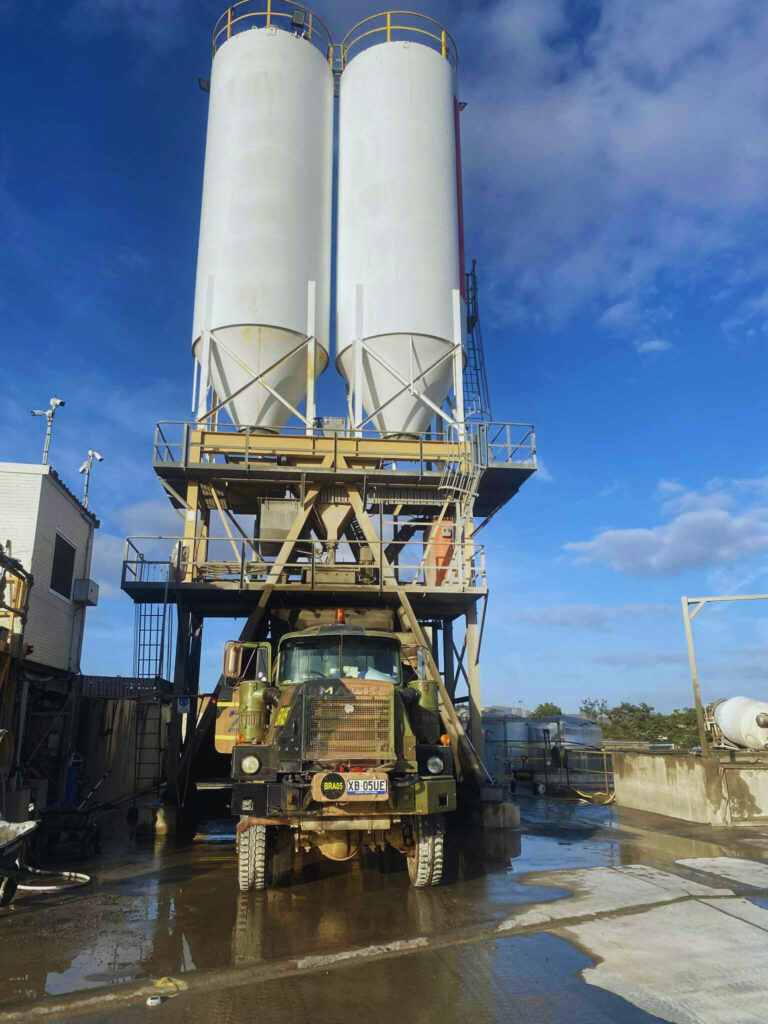 A large military truck is parked beneath two tall, white industrial silos on a concrete lot under a blue sky with some clouds. Scaffolding and machinery surround the silos, hinting at ongoing operations—learn more in the About BRAWG section.